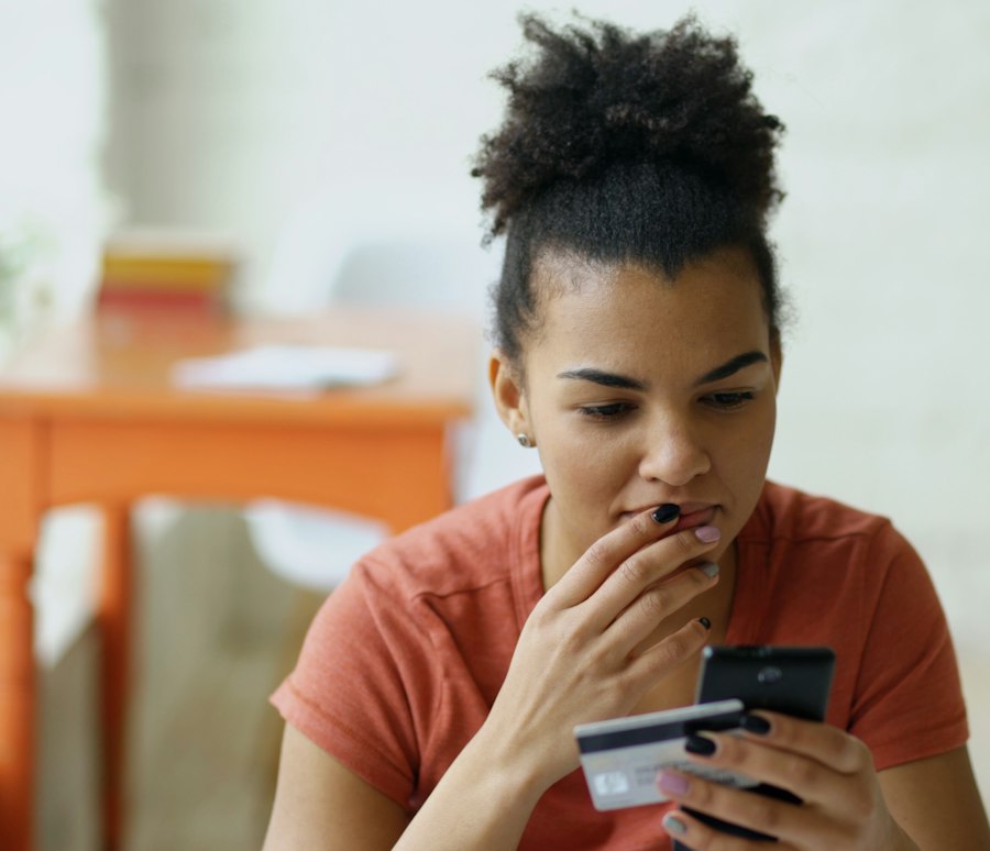 Mulher preocupada olhando o celular com um cartão de crédito na mão